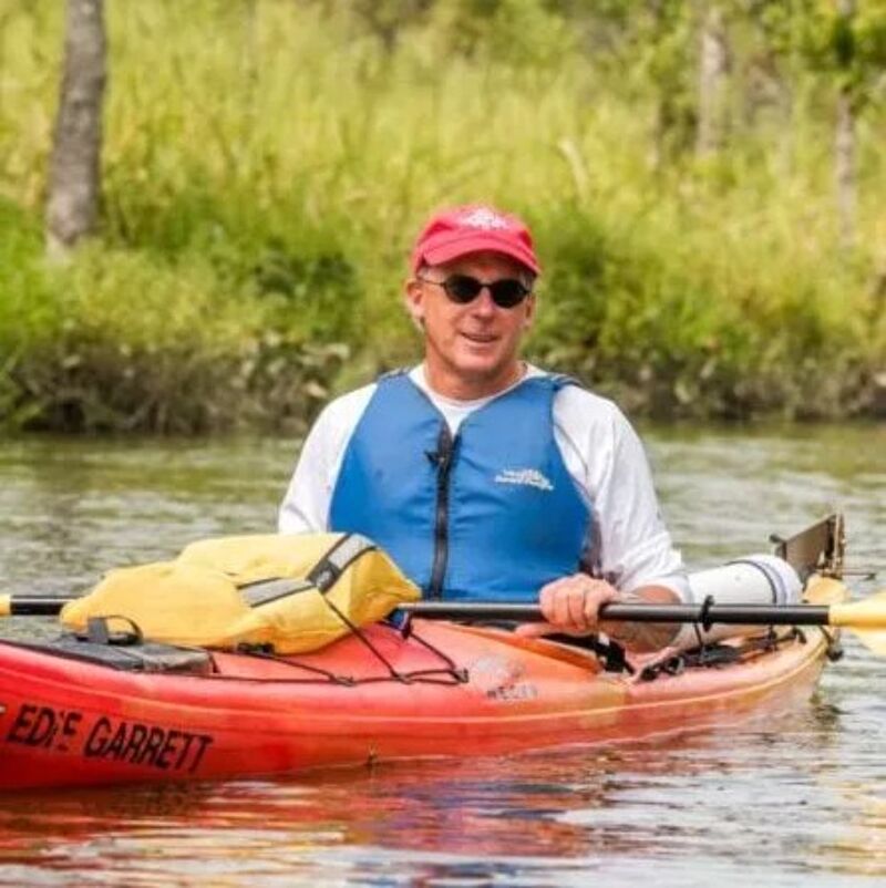 The image shows a man in a red kayak on a calm body of water. He is wearing a red cap, sunglasses, and a blue life vest. The kayak has the name "ED'S GARRETT" on it. The background features trees and greenery, suggesting a natural environment.